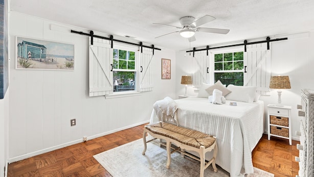 A bedroom with a bed, bedside tables, a bench, a ceiling fan, and two windows with shutters. - Cozy Holden Beach dog friendly beach cottage. Only 1.4 miles to Holden Beach on mainland side. (Supply)