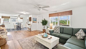 A spacious living room with a white coffee table, a green sofa, and a dining area with a white table and chairs. - Cozy Holden Beach dog friendly beach cottage. Only 1.4 miles to Holden Beach on mainland side. (Supply)