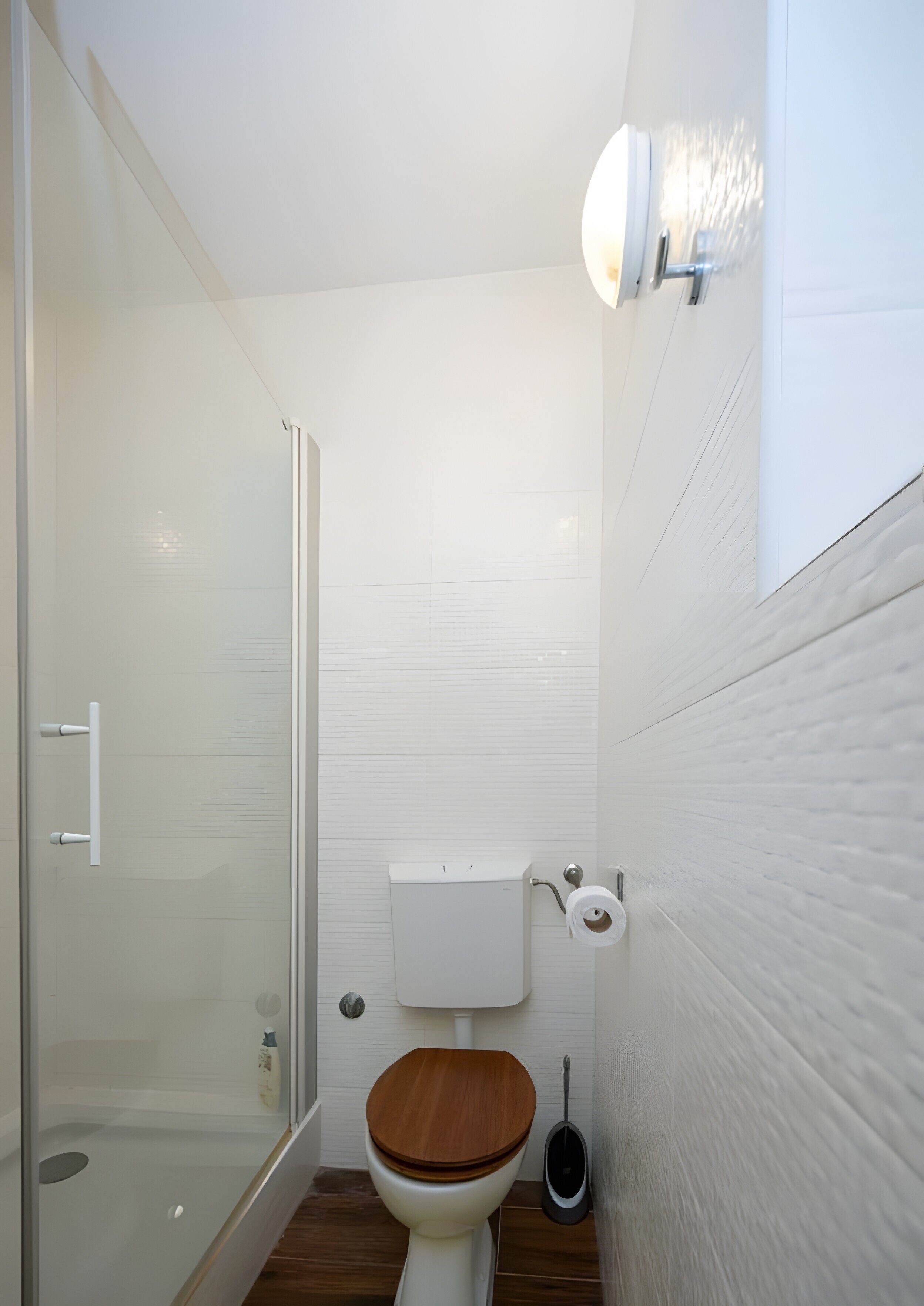 Une petite salle de bain moderne avec un abattant de toilettes en bois, une cabine de douche en verre et un mur recouvert de carreaux blancs.