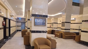 A lobby with a marble floor, beige walls, and a central column with a framed picture. There are several brown armchairs and a small table. - Qimmat AlAwali Hotel (Makkah)