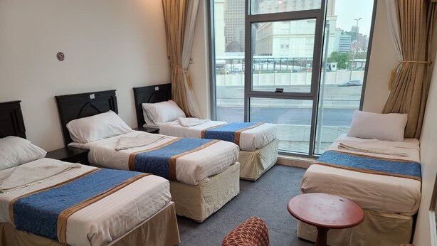 A hotel room with four beds arranged in a semi-circle, a small round table, and a patterned armchair. - Qimmat AlAwali Hotel (Makkah)