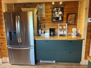 Une cuisine compacte équipée d’un réfrigérateur en acier inoxydable, d’une machine à café et d’un mur en bois orné d’une affiche sur le thème du café.