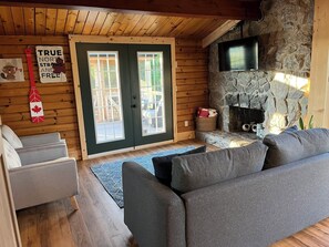 A cozy living room with a stone fireplace, a television mounted on the wall, and a large window with a view of the outdoors. - Serenity - Cottage on a private 4 acre waterfront lot surrounded by nature. (Kensington)
