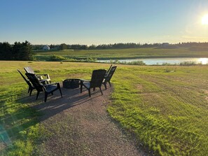 A gravel path leading to a fire pit with chairs, overlooking a body of water and fields. - Serenity - Cottage on a private 4 acre waterfront lot surrounded by nature. (Kensington)