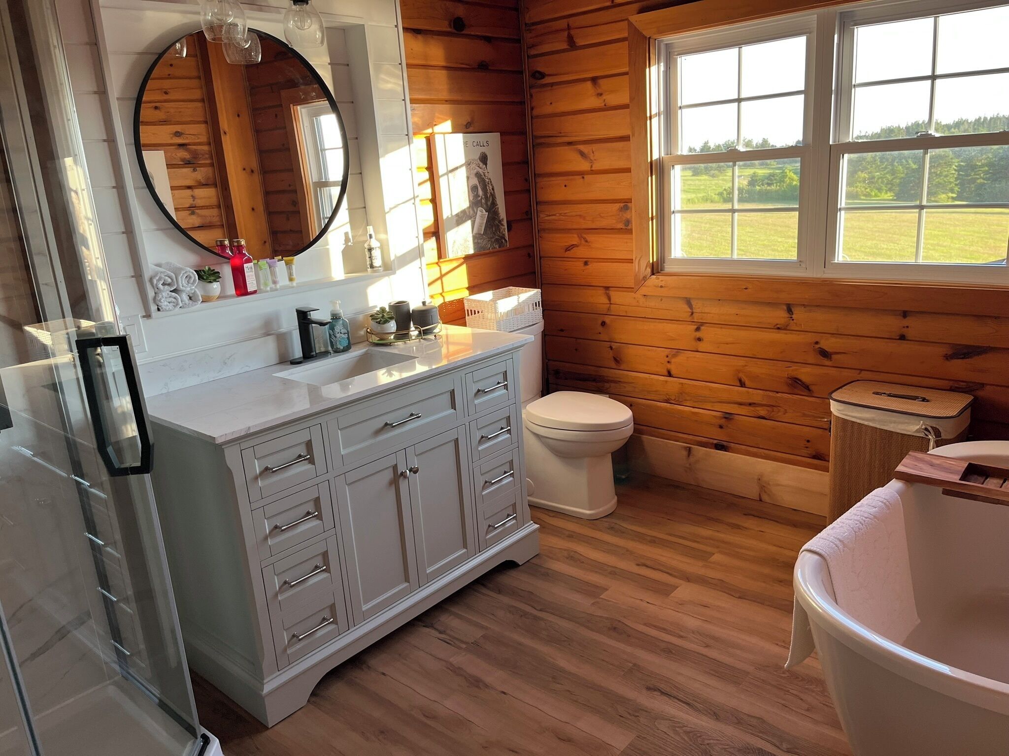A bathroom with a wooden interior, a white sink with multiple drawers, a toilet, and a window with a view of a green landscape.