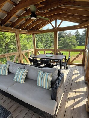 Un espace extérieur aménagé, abrité et doté d’un canapé, d’une table et de chaises, entouré de rampes en bois et offrant une vue sur la verdure.