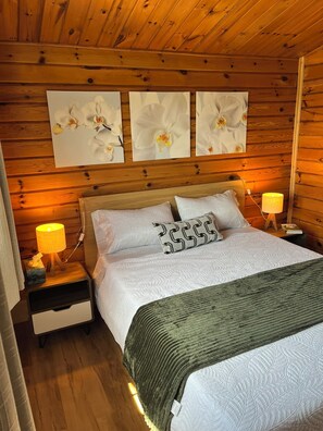 A wooden cabin bedroom with a bed, two bedside lamps, and three framed floral artworks on the wall. - Serenity - Cottage on a private 4 acre waterfront lot surrounded by nature. (Kensington)