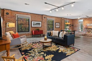 A spacious living room with a brick wall, a blue sofa, a wooden coffee table, and a floral rug. - Magnolia Market Loft | Steps to Old Town & Trails (Fort Collins)