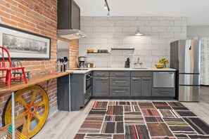 Une cuisine moderne avec un mur en briques, des armoires grises, un réfrigérateur en acier inoxydable et un tapis coloré.