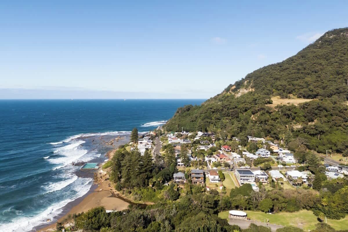 A coastal residential area with houses, a beach, and a hill with dense vegetation.