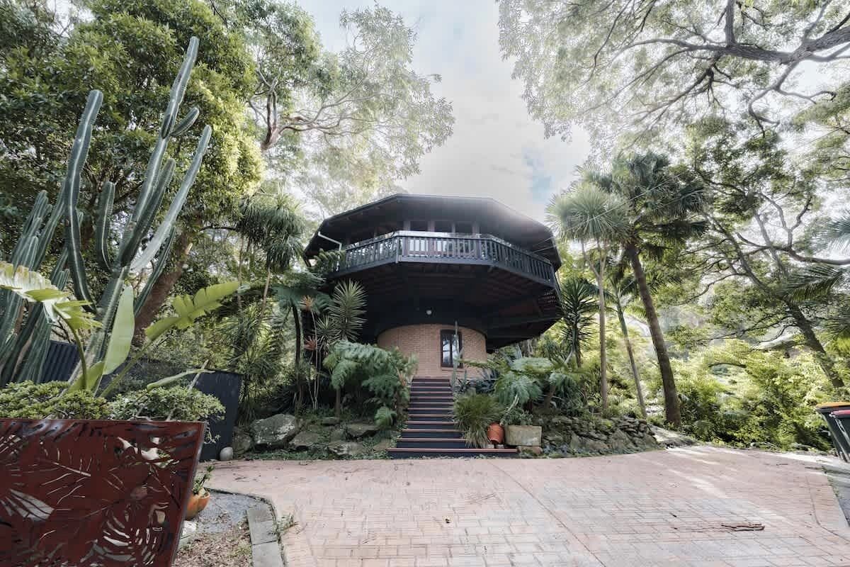 Un bâtiment circulaire doté d’un balcon, entouré d’une végétation luxuriante et accessible par un chemin pavé menant à l’entrée.