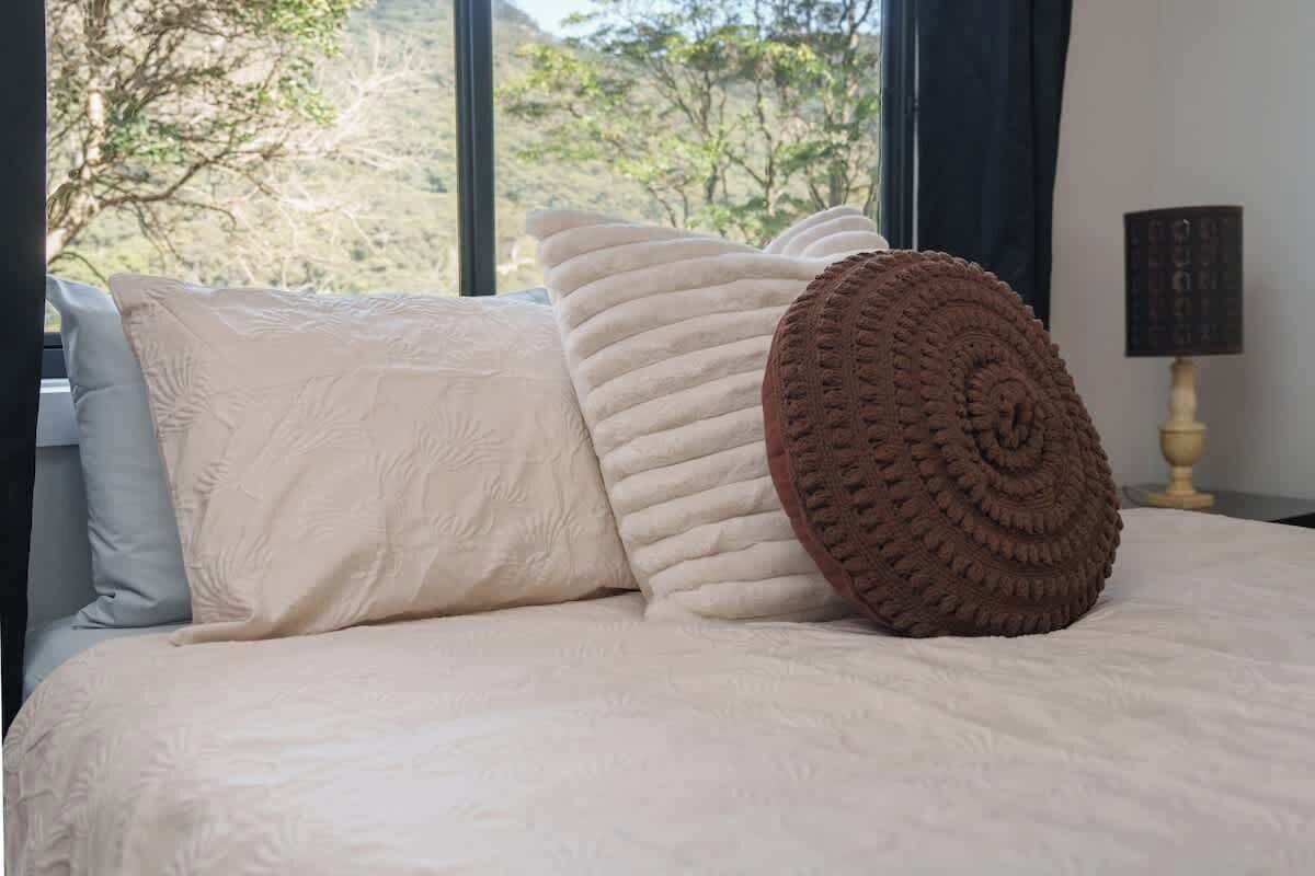 Un lit recouvert d’une couvre-lit beige, agrémenté d’un coussin décoratif marron, et une fenêtre donnant sur des arbres.