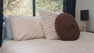 Un lit recouvert d’une couvre-lit beige, agrémenté d’un coussin décoratif marron, et une fenêtre donnant sur des arbres.