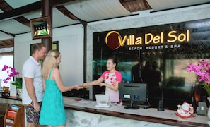 En receptionsdisk på Villa Del Sol Beach Resort & Spa, utrustad med en digital informationskiosk, ett inramat certifikat och en vas med blommor.