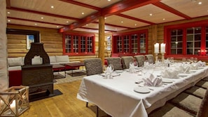 Una larga mesa de comedor dispuesta para una comida en una habitación de estilo cabaña de madera, con paredes de madera y ventanas con marcos rojos.