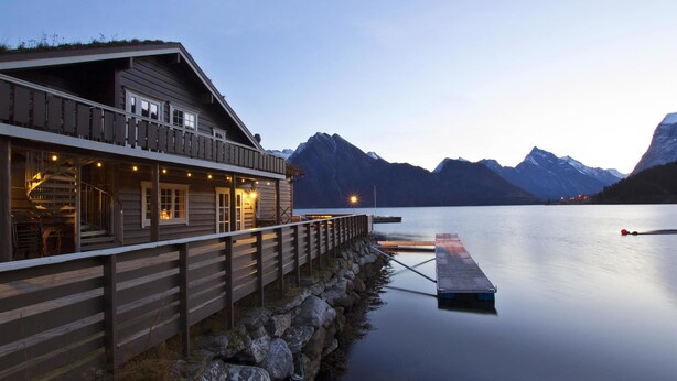 A wooden cabin with a balcony overlooking a calm lake, surrounded by mountains.