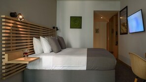 A modern bedroom with a wooden headboard, a bedside table with a glass of wine, a bed with white bedding, a green abstract painting, and a TV on the wall. - Fos Hotel (Athens)