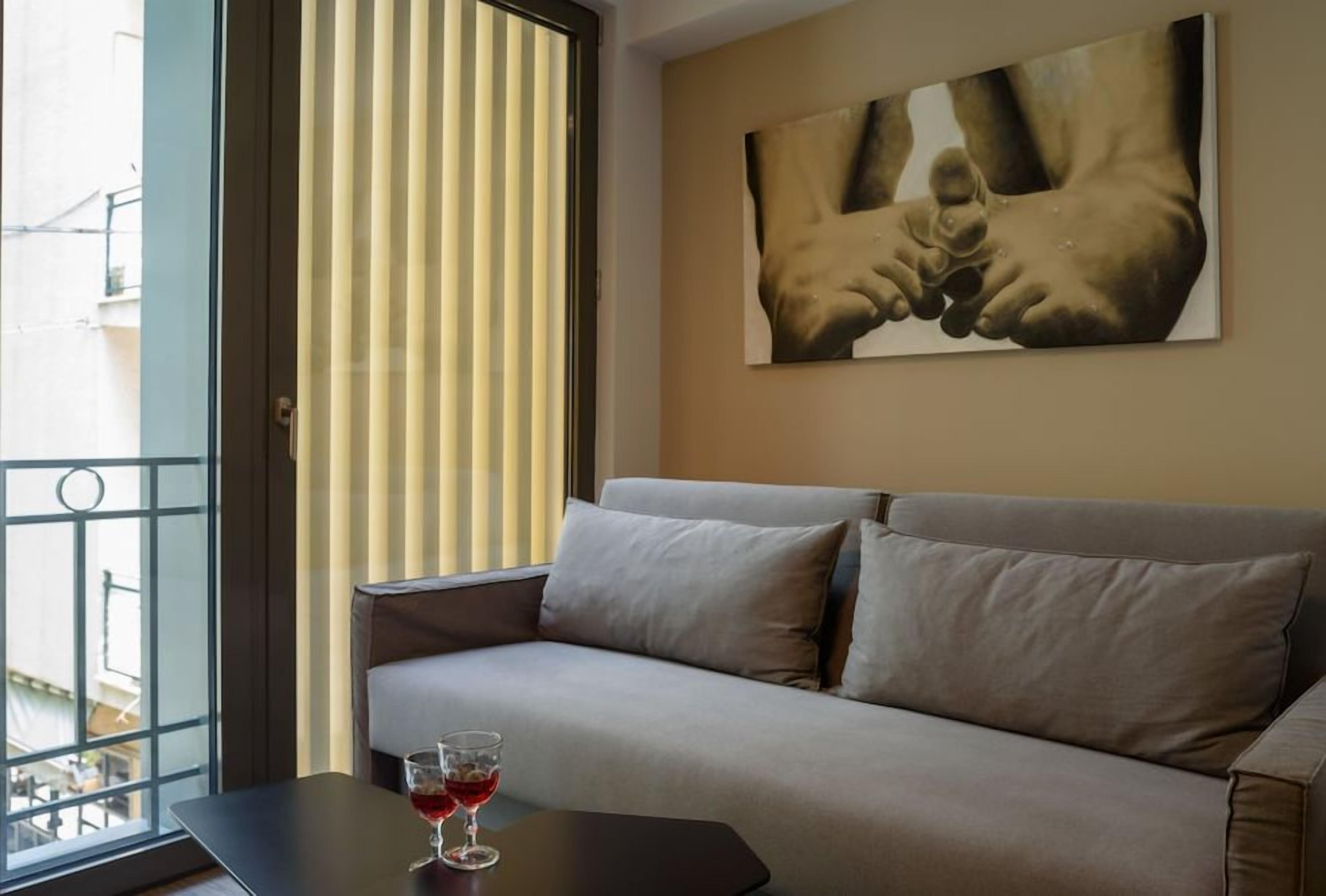 Una habitación con un sofá, una mesa de vidrio con una copa de vino, una ventana con persianas y un cuadro en la pared.