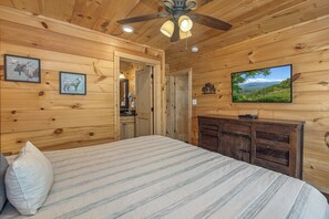 A wooden cabin bedroom with a bed, a ceiling fan, a mounted television, and a door leading to a bathroom. - Welcome to Sleepy Bear, a spacious three-bedroom, three-full-bath with a Swim Sp (Sevierville)