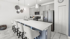 A modern kitchen with a central island, stainless steel appliances, pendant lighting, and a clock on the wall. - Pebble Beach Retreat (Kissimmee)