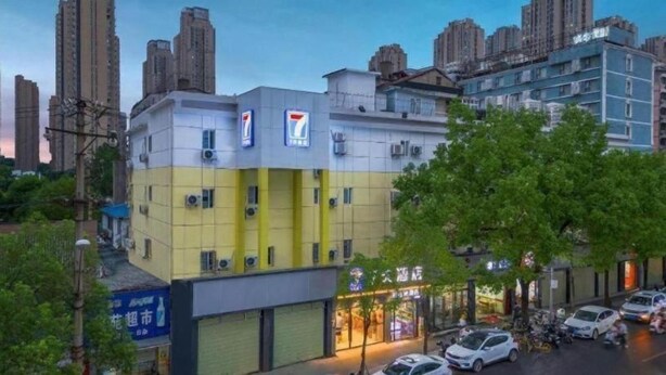 Un edificio de varios pisos con una tienda de conveniencia 7-Eleven en la planta baja, rodeado de coches estacionados y árboles.