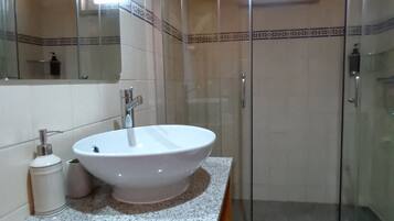 Un baño con lavamanos blanco, espejo, cabina de ducha y tocador de madera.