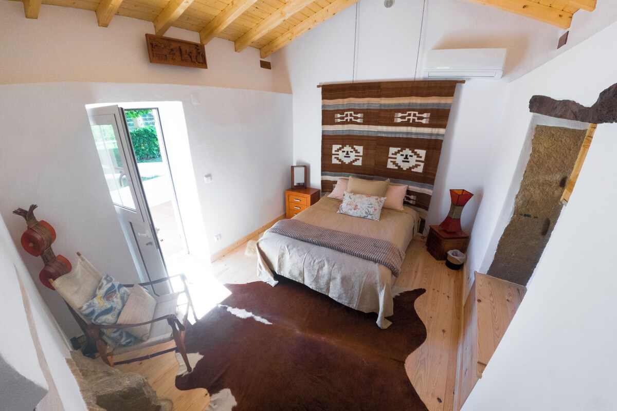 Un dormitorio con cama, techo de madera, ventana, silla y alfombra de cuero de vaca.