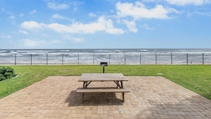 A picnic table on a brick patio with a view of the ocean and a metal fence. - Las Brisas 523 (New Smyrna Beach)
