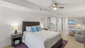 A bedroom with a bed, bedside tables, a ceiling fan, and a view of the bathroom and kitchen. - Las Brisas 523 (New Smyrna Beach)