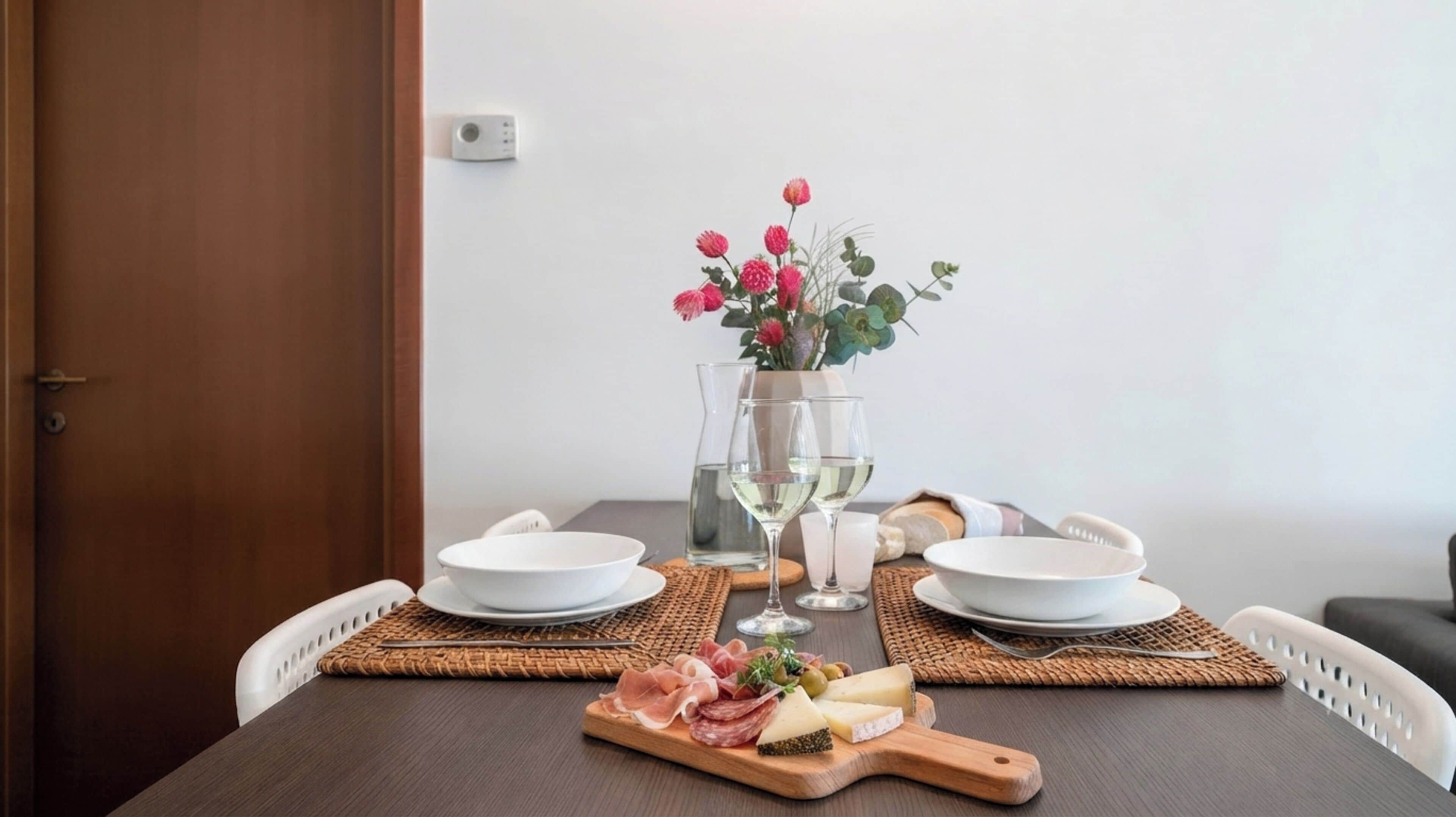 A dining table with a white plate, a glass of water, a glass of wine, a wooden board with cheese and cured meats, and a vase with pink flowers.