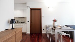 A modern kitchen with white cabinets, a wooden door, and a dining area with a table and chairs. - Italianway Comfort - Vico Magistretti 1 B (Milan)