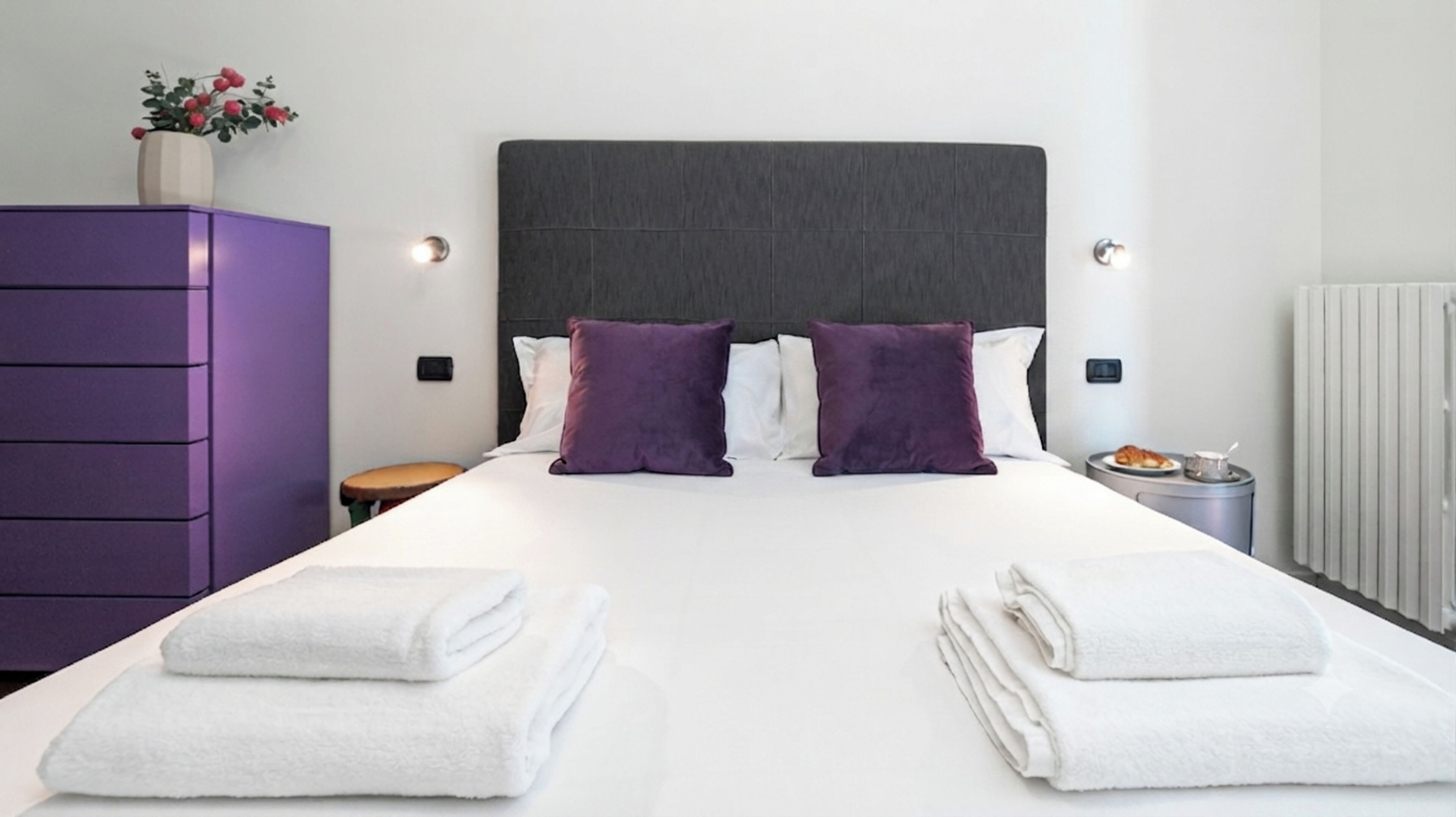 A hotel room with a large bed, two pillows, a dresser, a small table, and a radiator.