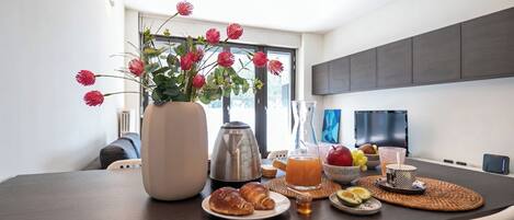 Una mesa de comedor con un florero, una cafetera, croissants, un frutero y un vaso de jugo de naranja.
