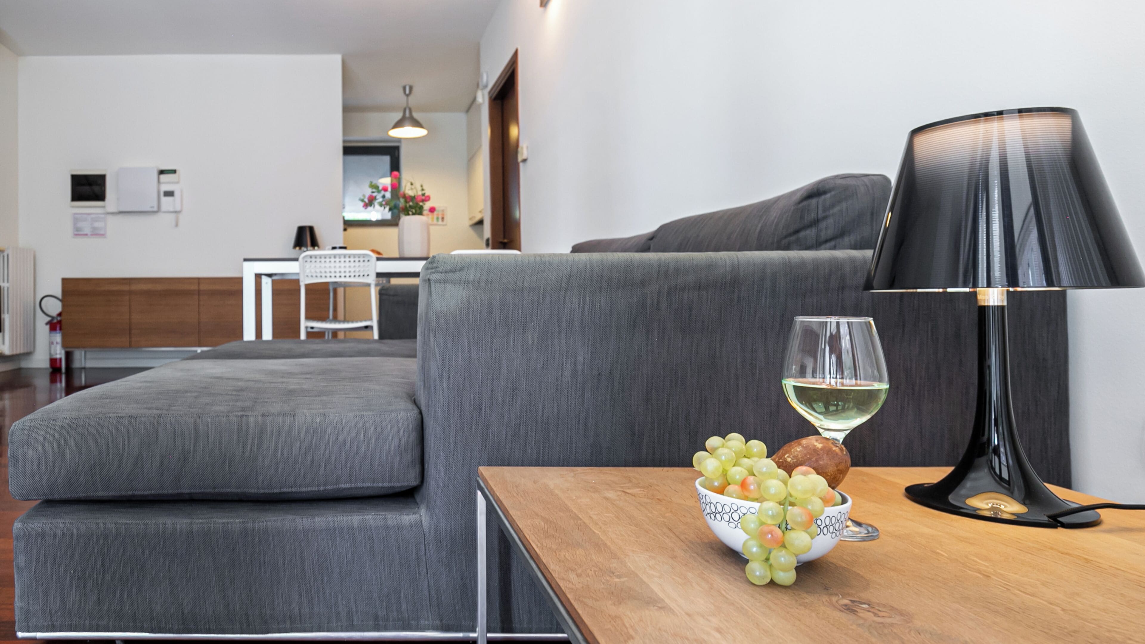 A modern living room with a grey sofa, a wooden coffee table with a bowl of grapes and a wine glass, and a black floor lamp.