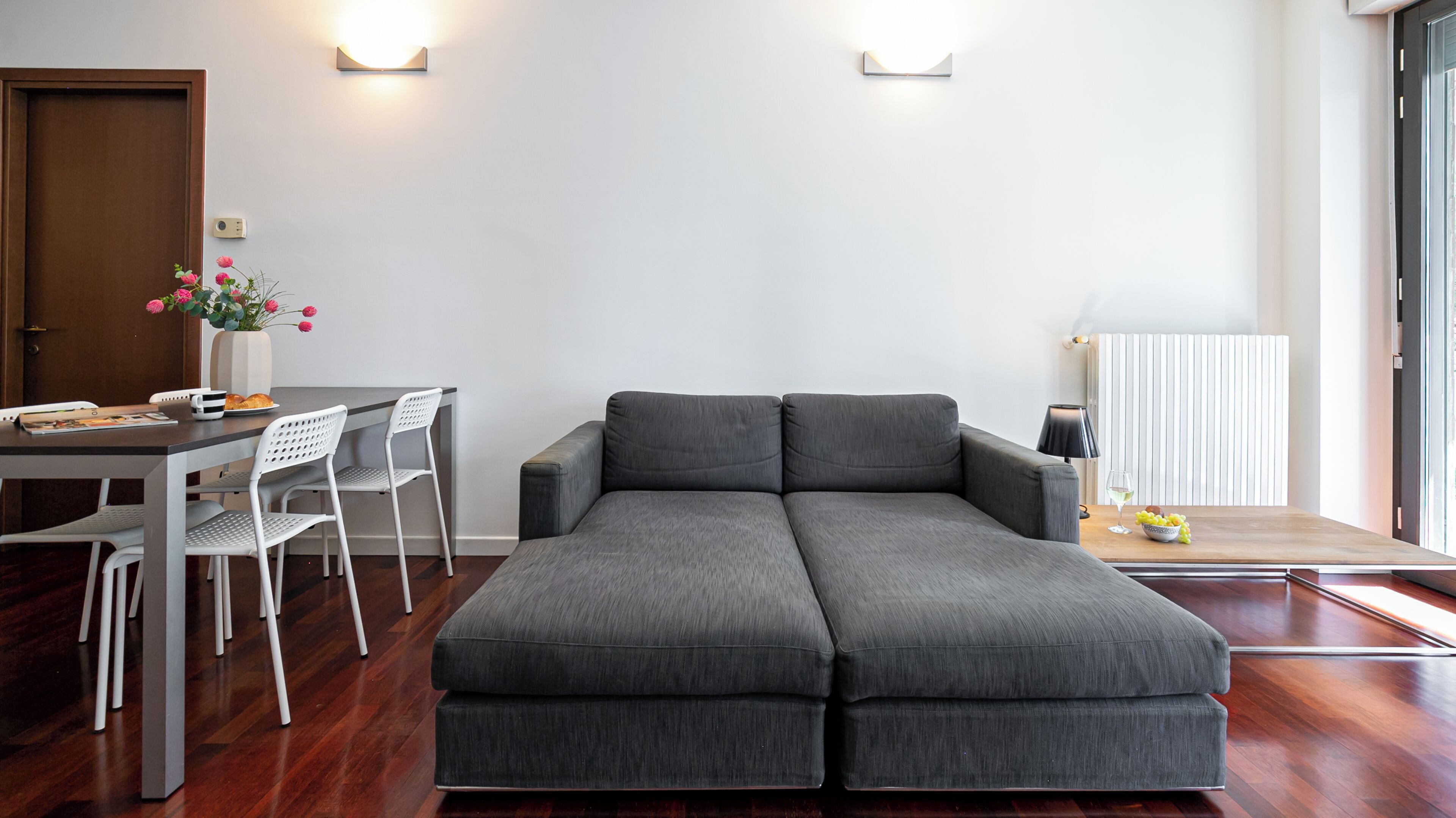 A modern living room with a grey sectional sofa, a dining table with chairs, a vase with flowers, and a lamp on a side table.