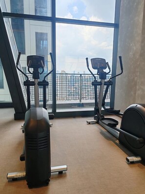 A gym with two exercise bikes and a stationary bike, large windows with a city view, and a carpeted floor. - Spire Skyline Suite | Luxury 45 sqm + Pool, Gym, and Netflix (Makati City)