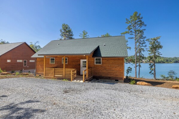 A wooden cabin with a gravel driveway, a small porch, and a view of a lake.