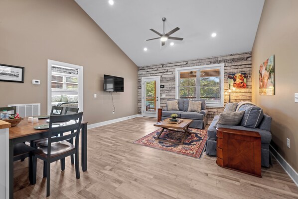 A spacious living room with a high ceiling, a ceiling fan, a large window, a fireplace, and a dining area with a table and chairs.