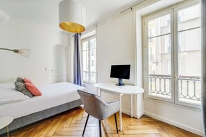 A hotel room with a bed, a desk with a computer, a chair, and large windows. - Charming Appartement 1ch/4p - Paris 17e (Paris)