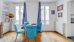 A room with a round glass table, two blue chairs, a tripod floor lamp, and a radiator. - Charming Appartement 1ch/4p - Paris 17e (Paris)