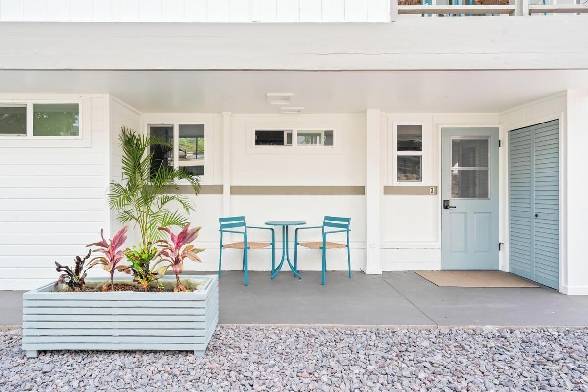 A white building with a balcony, a planter with plants, two tables with chairs, and a door with a number.