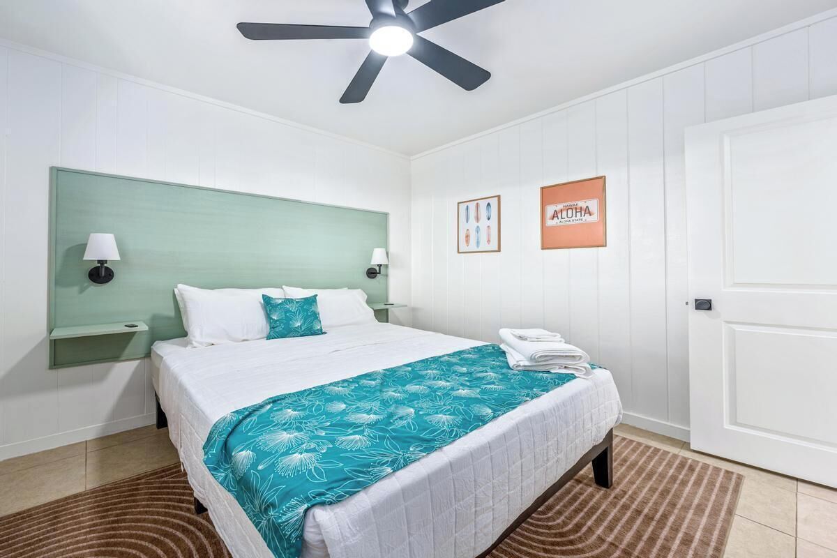 A hotel room with a bed, a ceiling fan, a nightstand, wall art, and a door.