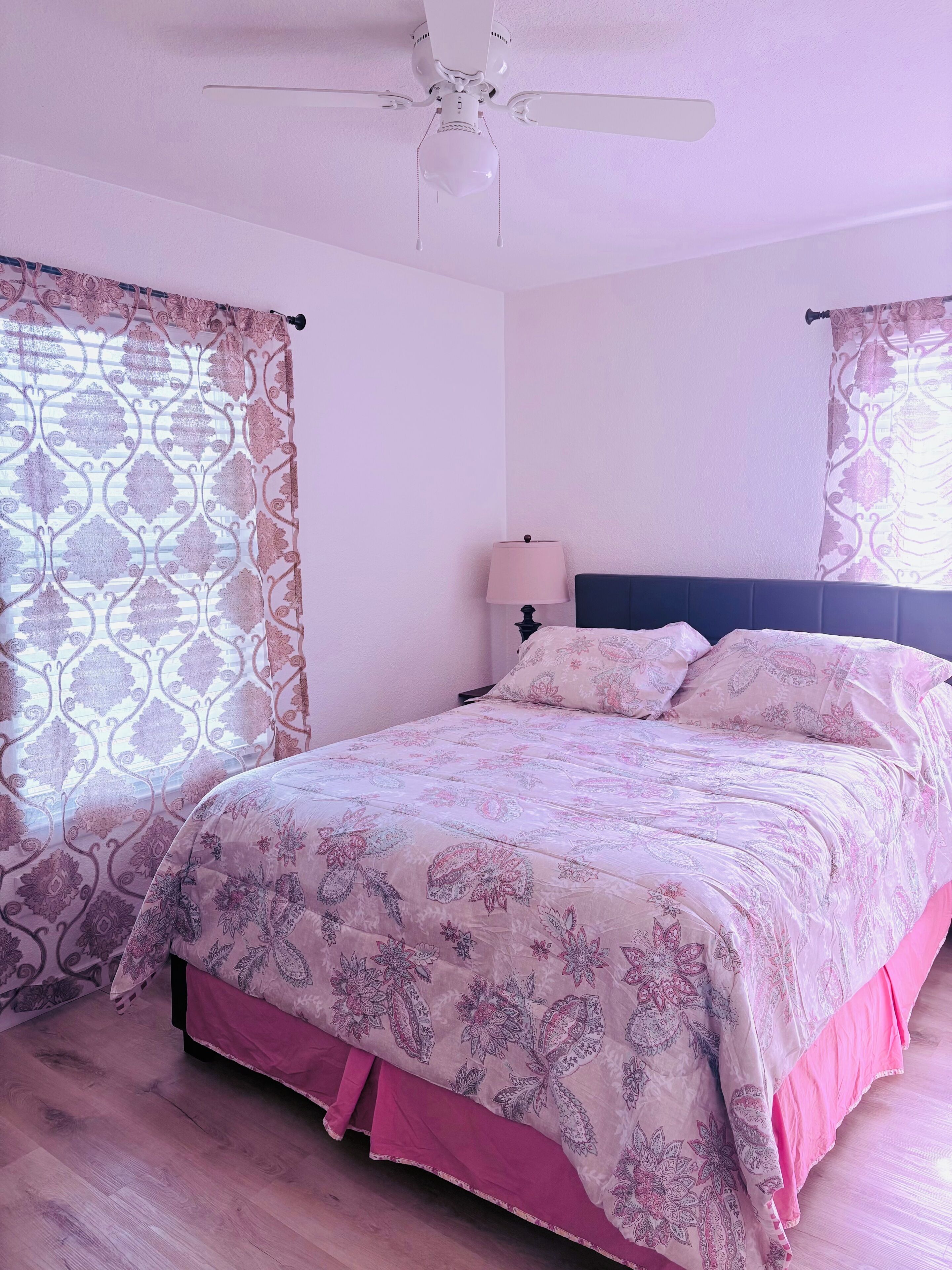 A bedroom with a bed, curtains, a ceiling fan, and a bedside lamp.