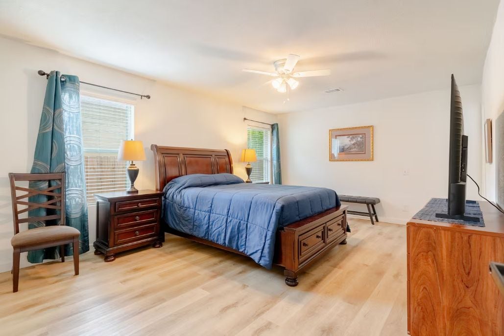 A bedroom with a bed, a dresser, a chair, a desk, a ceiling fan, and a television.