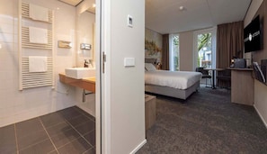 Een moderne hotelkamer met een bed, bureau, televisie en een badkamer met handdoekhouder en wastafel.