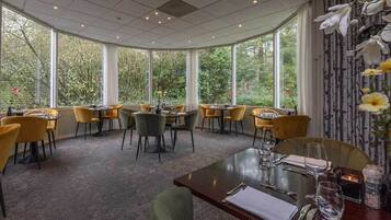 A restaurant with round tables and chairs, large windows with a view of trees, and a carpeted floor.