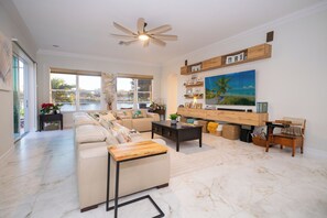 A spacious living room with a large sofa, a coffee table, and a TV mounted on the wall. There are windows providing natural light and a view of the outdoors. - The Ultimate Waterfront Retreat | 5BR • Pool & Spa • Resort Amenities (Cape Coral)