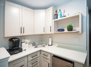Uma cozinha moderna com armários brancos, uma cafeteira, uma pia e uma estante com garrafas e uma planta.