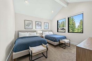 Ein Schlafzimmer mit zwei Betten, einem kleinen Tisch und einer Bank. An der Wand befinden sich gerahmte Pflanzenbilder und ein Fenster mit Blick auf Bäume.