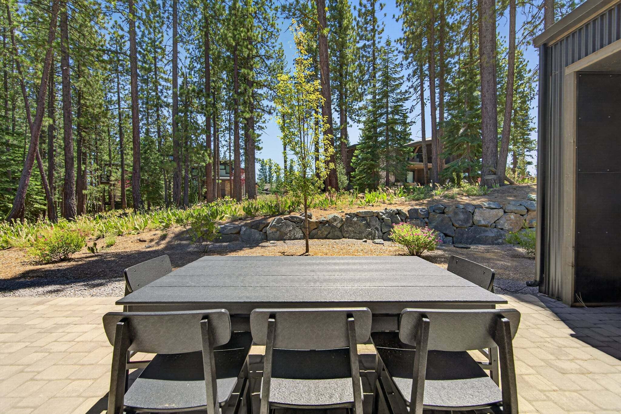 Un patio al aire libre con una mesa rectangular y seis sillas, rodeado por un muro de piedra y varios árboles.